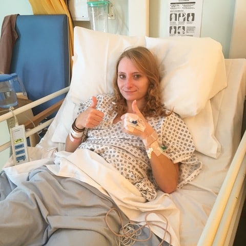 Charlotte, a white woman with long blonde hair, is lying in a hospital bed with her thumbs up.