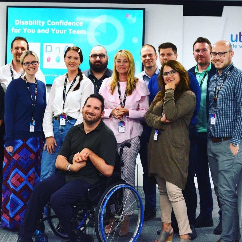 A group of people standing or sitting in front of a presentation board that reads: Disability confidence for you and your team. Everyone is looking at the camera and smiling.