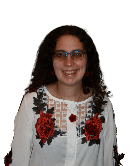 ropped head shot of a white woman with shoulder length brown curly hair, and brown eyes. She is wearing blue & pink glasses, and a white shirt with red roses, with matching rose necklace and earrings. She looks at the camera and smiles. She is indoors and standing before two framed paintings featuring ancient Egyptian imagery