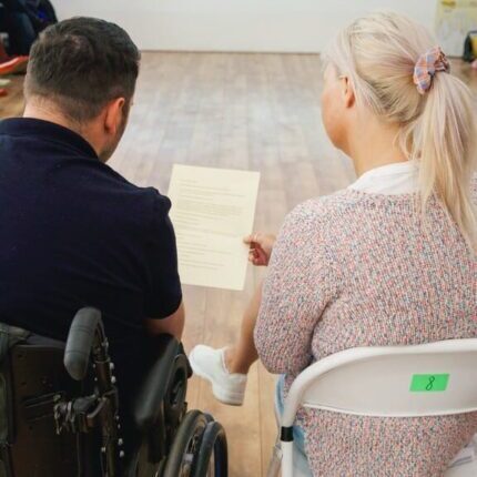 A wheelchair user is looking at a piece of paper held by his personal assistant.