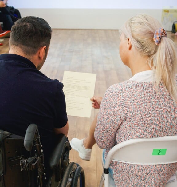A man in a wheel chair and a woman on a chair look at a piece of paper.