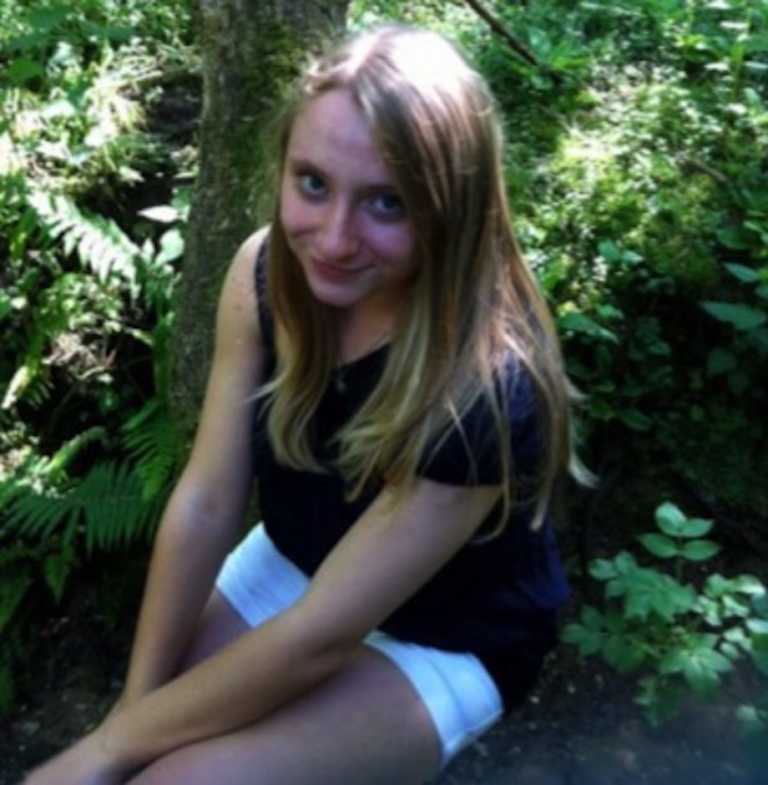 Charlotte, a young white girl with blonde hair, sitting by a tree and smiling at the camera.