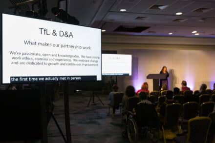 In the foreground a white screen with text reads; TfL and D&A What makes our partnership work We're passionate, open and knowledgeable. We have strong work ethics, stamina and experience. We embrace change and are dedicated to growth and continuous improvement. In the background, unfocused, is Venetia Petter, presenting to a conference. 