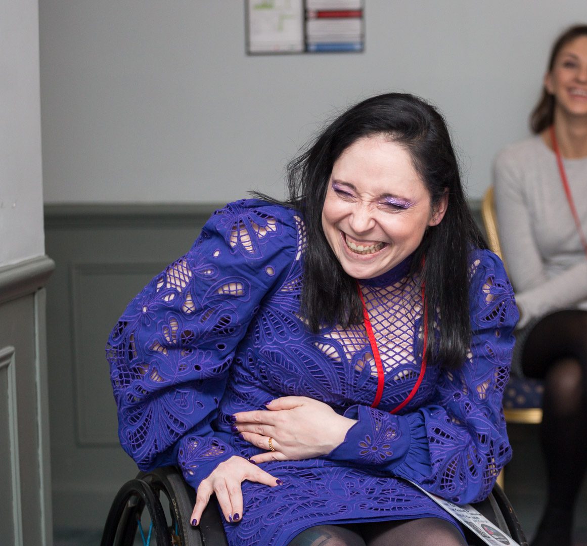 A non-binary wheelchair user wearing a purple dress is smiling in a training session.