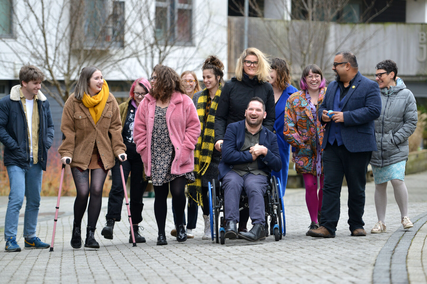 12 members of the D&A team are outside. They are in movement towards the camera, but as they move are looking at each other and laughing or smiling. They are a diverse group with different disabilities, races, genders and heritages.