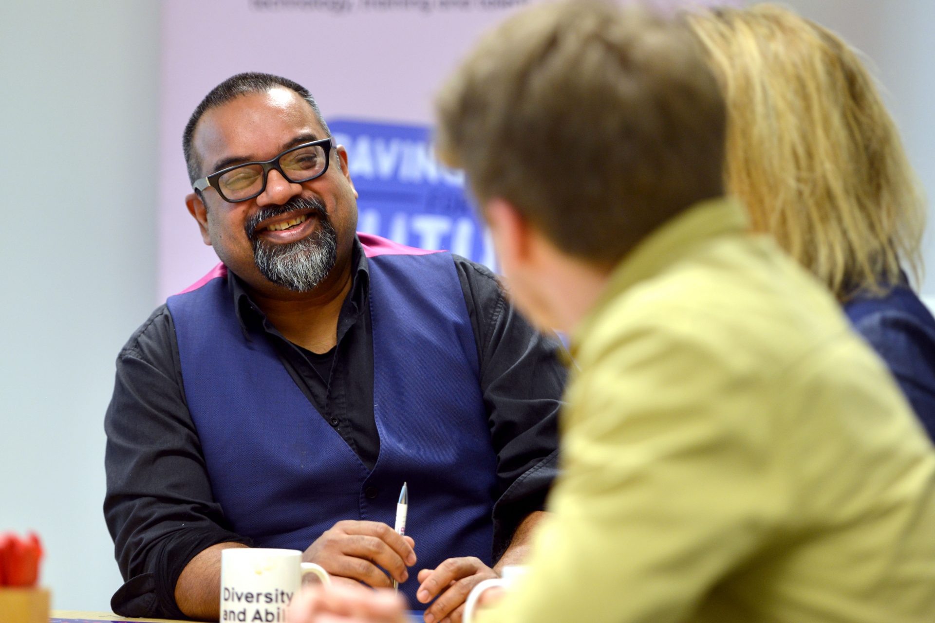 In an office, three people are sat around a table. In focus is Atif, who is smiling at two people out of focus in the foreground.