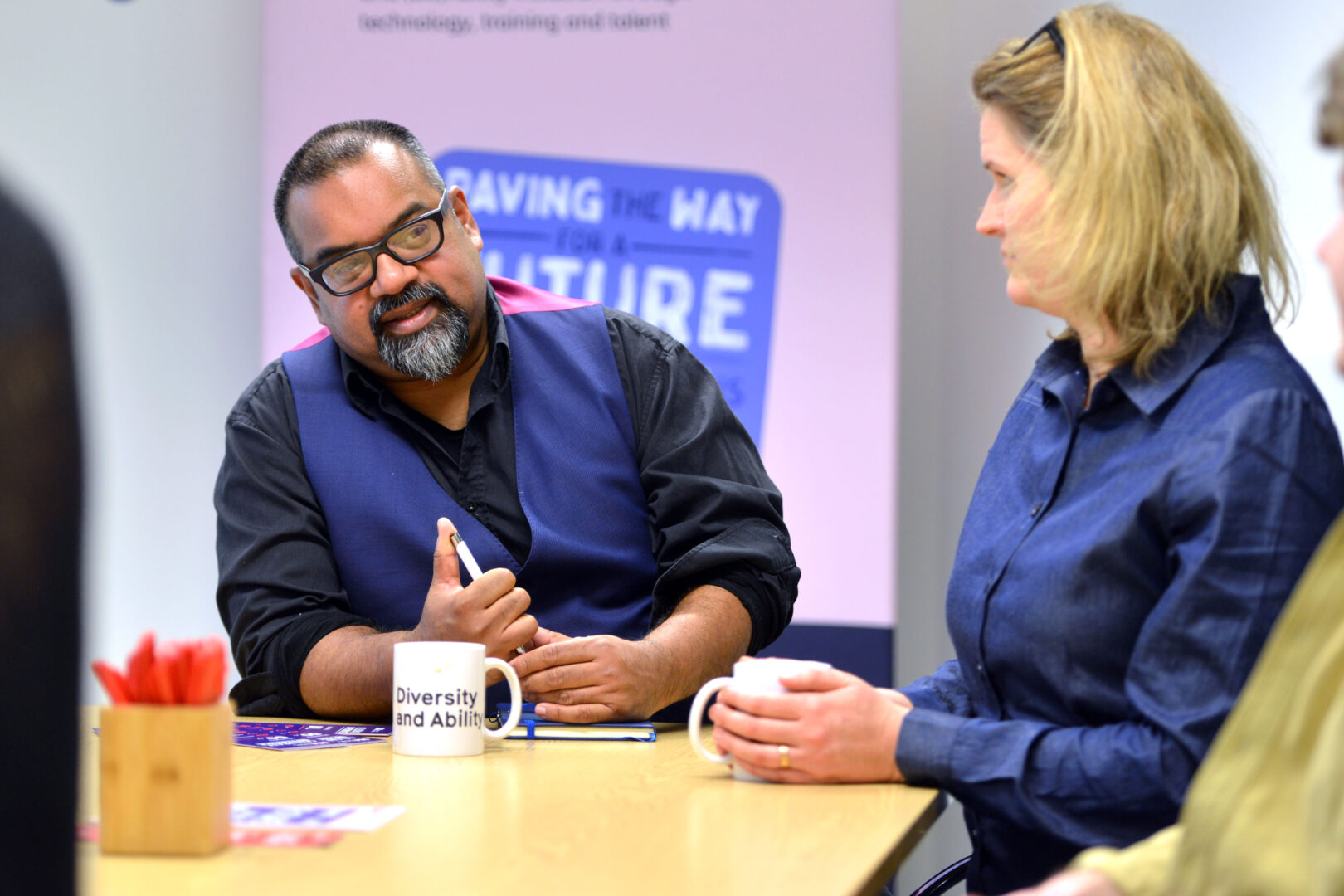 Members of the D&A team around a table in the office. In focus is Atif, a brown man wearing glasses, a black shirt and blue waistcoat. He is speaking and looking intently at Giedre, a white woman with blonde straight hair, who is sat to his right.