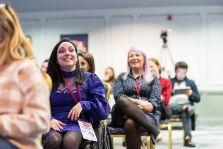 A wheelchair user is smiling in the audience of a conference presentation.