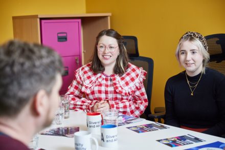 Three people are having a meeting. A man has his back to the camera and two women listen to him smiling.