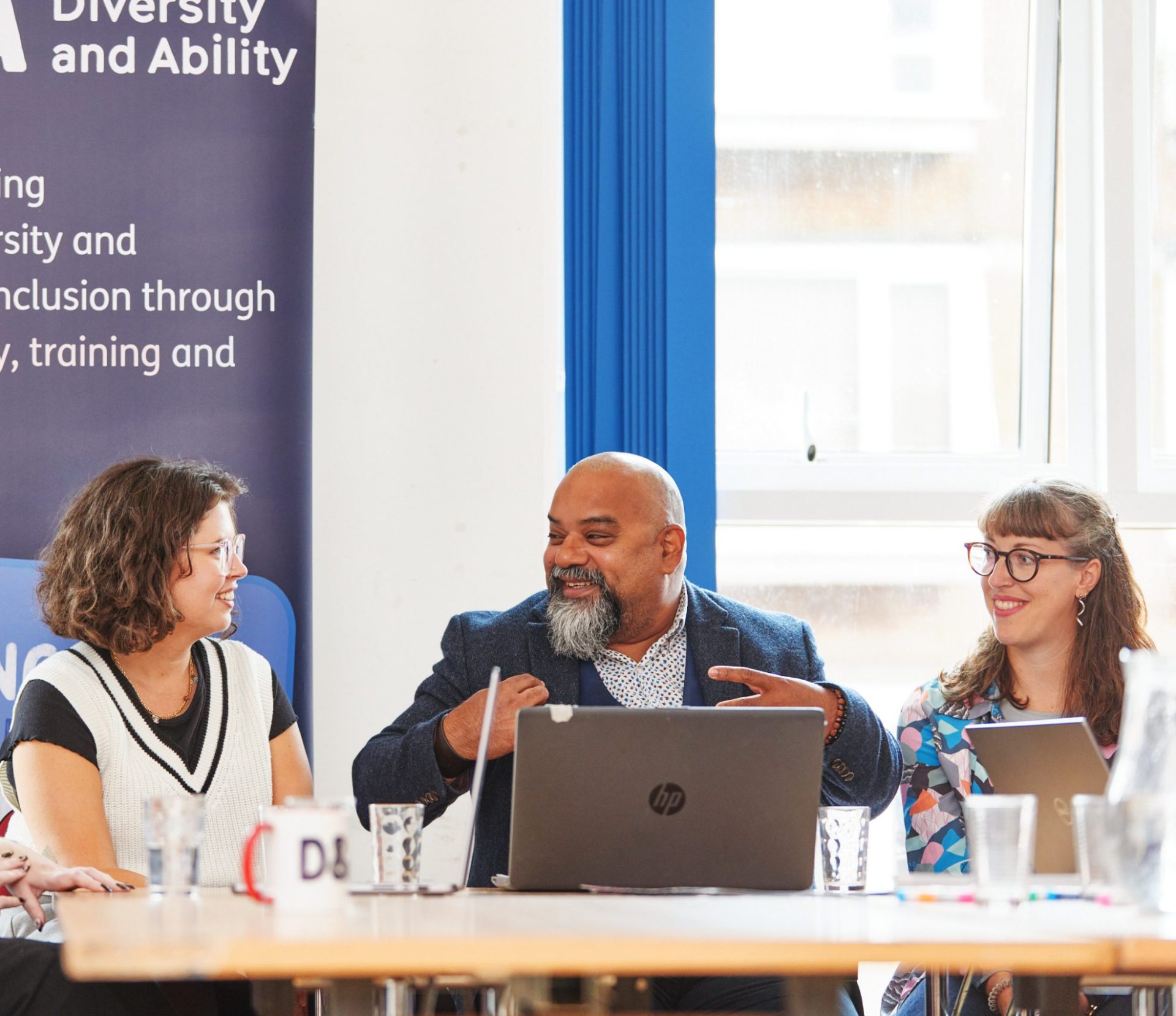 A group of people sat around a table. In the centre is a brown man with a grey beard, who is smiling as he speaks, gesturing with his hands. Either side of him are two white women, both wearing glasses, smiling at him as they listen.