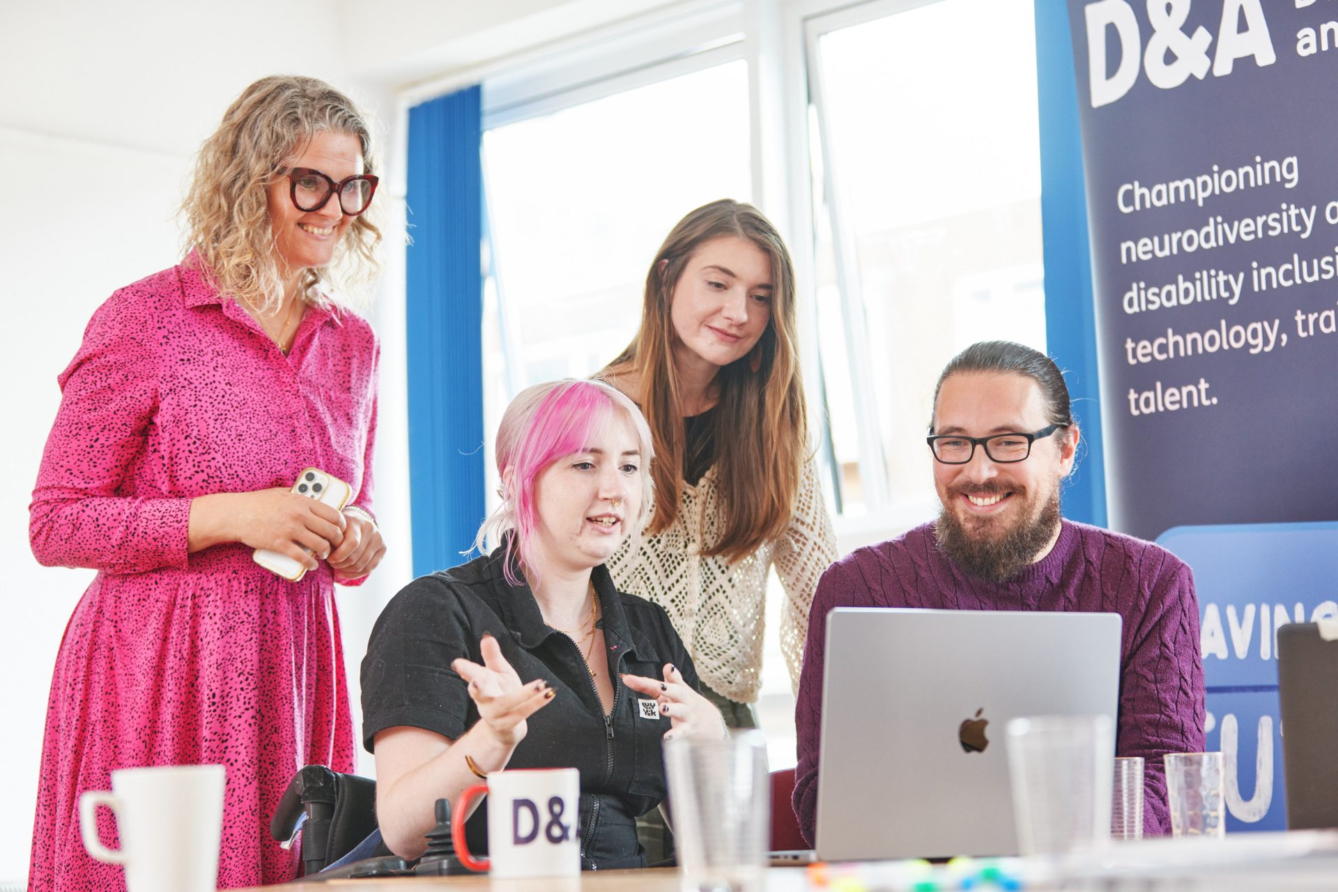 Four people are clustered around a laptop. One, a white woman in a wheelchair with pink hair, is speaking, using her hands to gesture. The other three are smiling at the screen as she speaks.