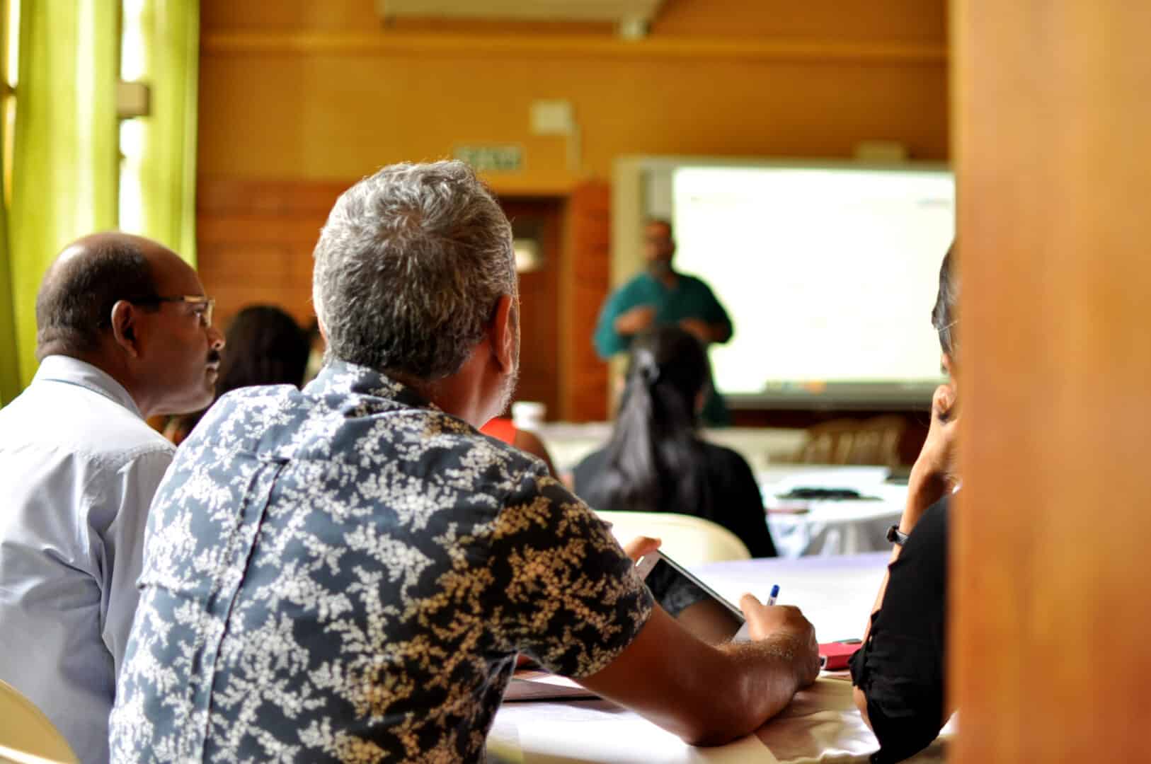 In a classroom, a dark-skinned person with short grey hair has their back to the camera. They are facing a person at the fair end of the room who is standing in front of a screen and speaking.