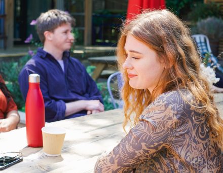 a white woman with strawberry blonde hair sits on a picnic bench in the sunshine. 