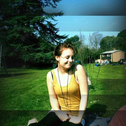 A younger Brigit, a white girl with brown hair, sits on a grass lawn in the sun. 