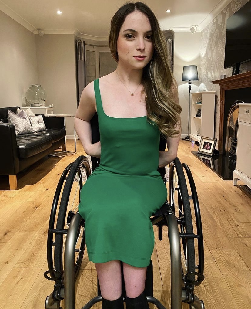 Caitlyn, a young white woman with blonde hair, is wearing a bright green dress and sitting in her wheelchair. Her hair is swept to one side and her hands are on her hips. She is smiling slightly at the camera.