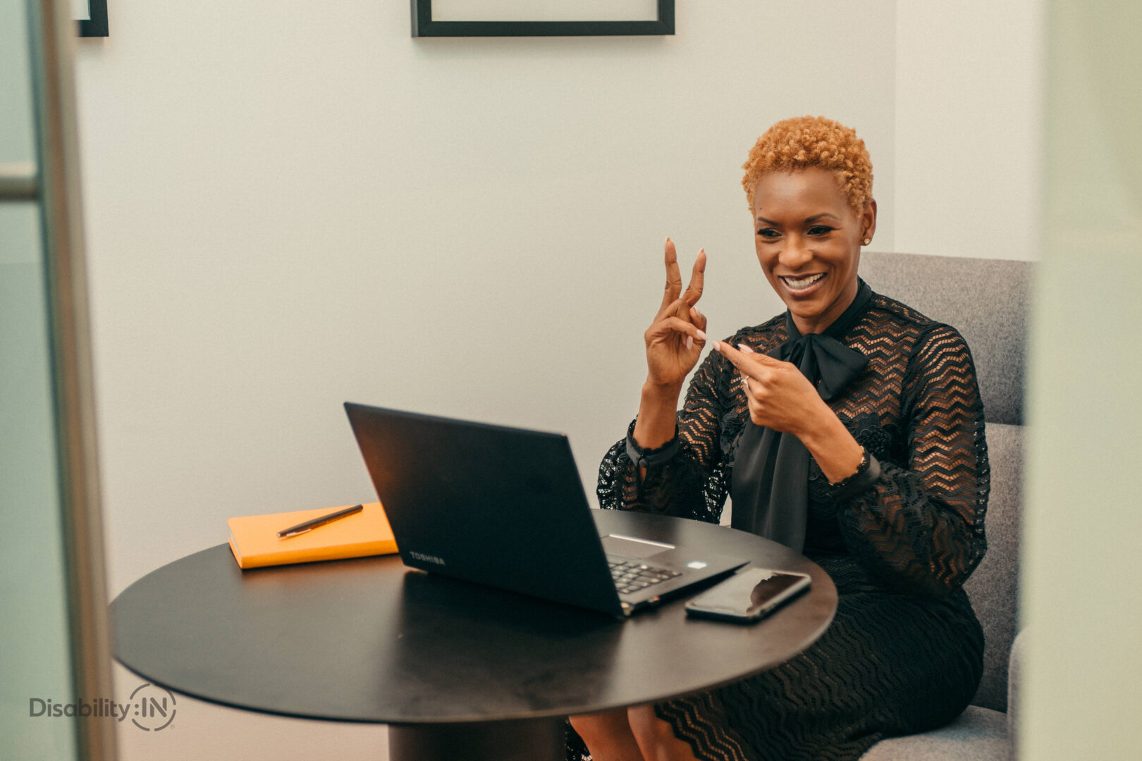 A woman of colour smiles and signs in American Sign Language to a laptop as if it's a video meeting.