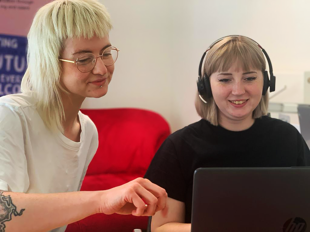 Two people sit at a desk in front of a laptop screen. The person on the left is a non-binary white person, who is pointing at the laptop screen as if demonstrating something. The person on the right is a white woman, who is wearing headphones and smiling as she focuses on the laptop screen.