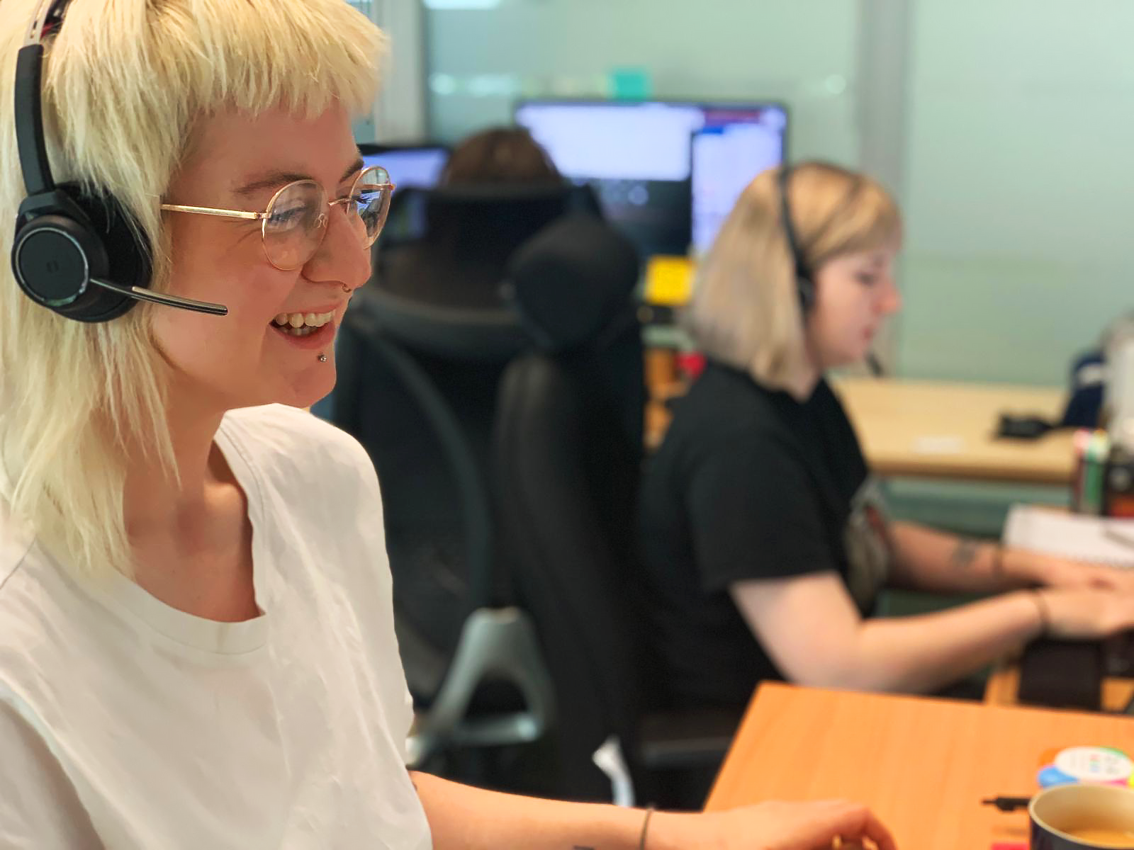 A white person in an office is using a noise cancelling headset and microphone, smiling at their computer.