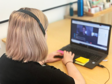 A woman with pink tinted hair is sat in front of a laptop in an online meeting.