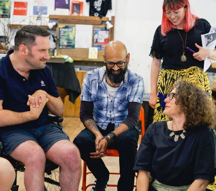 A wheelchair user, man on a chair, and a woman on the floor have a conversation at a training session.