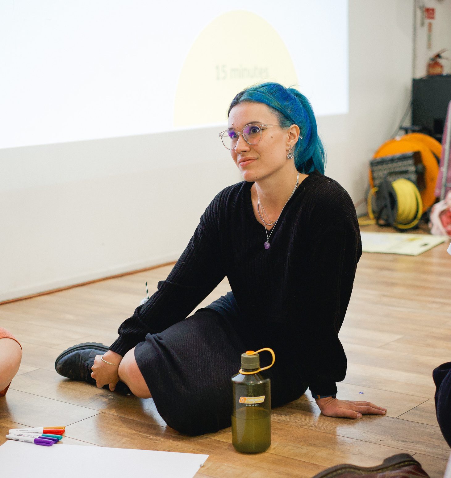 A white person sitting on the floor in a training session with glasses and black and blue hair.
