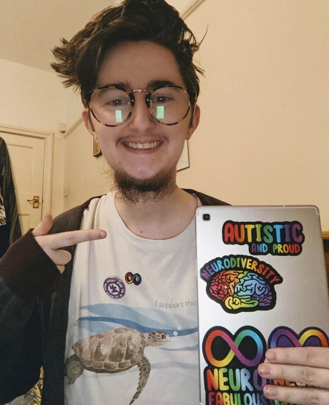 A white man is smiling at the camera. He has brown wavy hair and round brown glasses. He is wearing a cardigan over a white t-shirt with a turtle on it, with a rainbow autism infinity pin, and a purple badge that reads ‘autism speaks does not speak for me’. He is pointing to the back of a tablet he is holding, which has rainbow coloured stickers that read ‘autistic and proud’ and ‘neurodiversity’ above a rainbow brain graphic.
