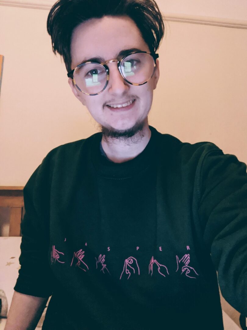 Jasper, a white man with short brown hair, a beard, moustache and glasses is smiling at the camera. He is wearing a green jumper with his name Jasper written out and the BSL sign for each letter illustrated below. 