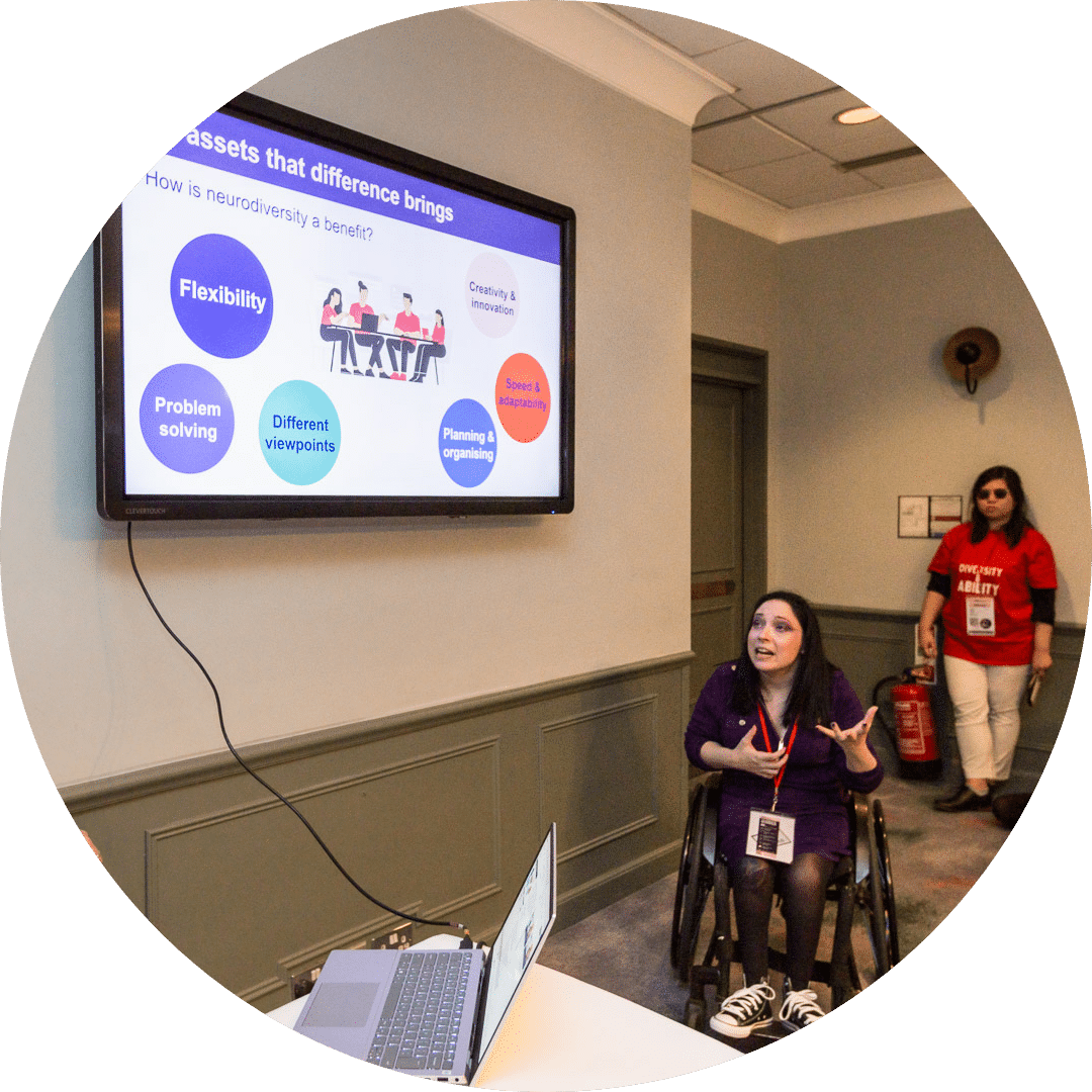 Rox, a person with long dark hair and wearing a purple dress, sits in their wheelchair as they present a slide from a wall-mounted tv screen. 