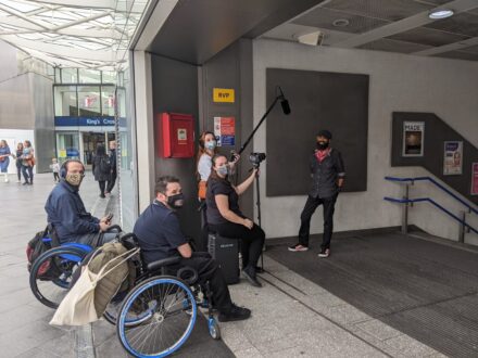 A group of 5 people, two in wheelchairs and the rest standing, in kings cross station filming a video. The team are all wearing masks and smiling at the camera.