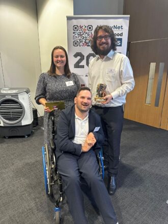 Adam, a wheelchair user, Meg and Billy stand smiling at the camera holding the tech4good awards trophies. 
