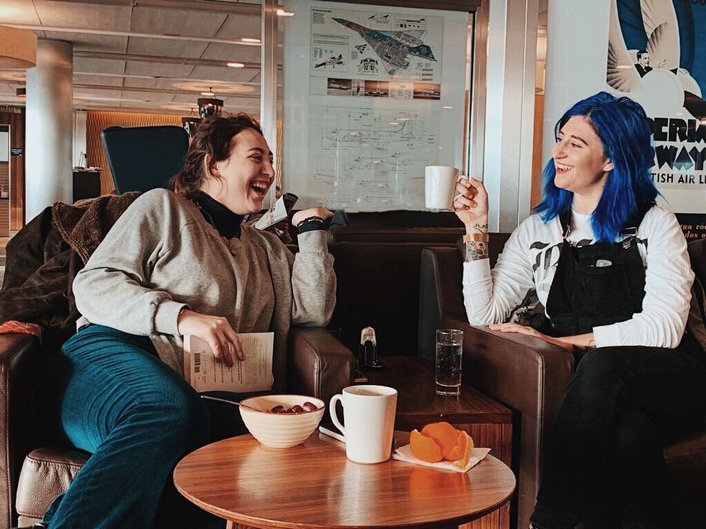 Nikita, a white, Jewish woman with curly brown hair is sitting with a closed book in her hand and laughing while facing her sister Tash. Tash is a white, Jewish woman with blue hair and tattoos on her arm. She is sitting with a mug in her hand, smiling and looking at her sister. Between them is a small table with a mug, cereal bowl and orange peels on it. 