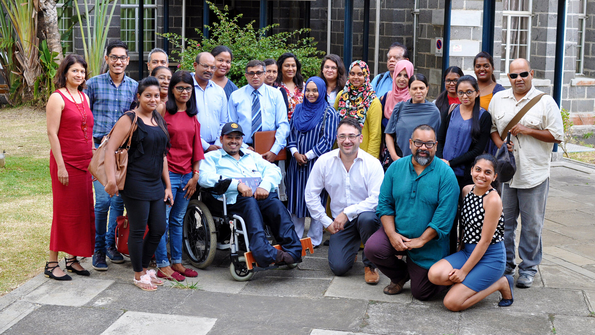 A group of people standing, sitting in wheelchairs or kneeling while smiling at the camera. They are a mixture of different races, skin colours, genders, ages and disabilities. They are in a bright, outdoor setting in front of a modern bricked building.Amongst them is Atif, CEO of Diversity and Ability, a brown man with short dark hair and glasses. 