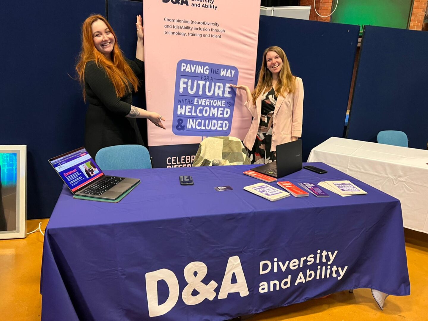 Two people are stood behind a D&A branded table. On the left is Alex, a white non-binary person with straight ginger hair, and on the left is Alice, a white woman with brown hair. Both are smiling and gesturing towards a banner that stands between them and reads: "D&A: paving the way for a future where everyone is welcomed and included"