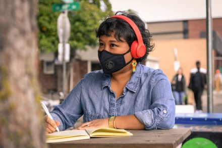 Close-up of a Filipinx woman with a filtering face mask, sitting at a table with a notebook and pen. She has colourful flower earrings and headphones on while looking into the distance.