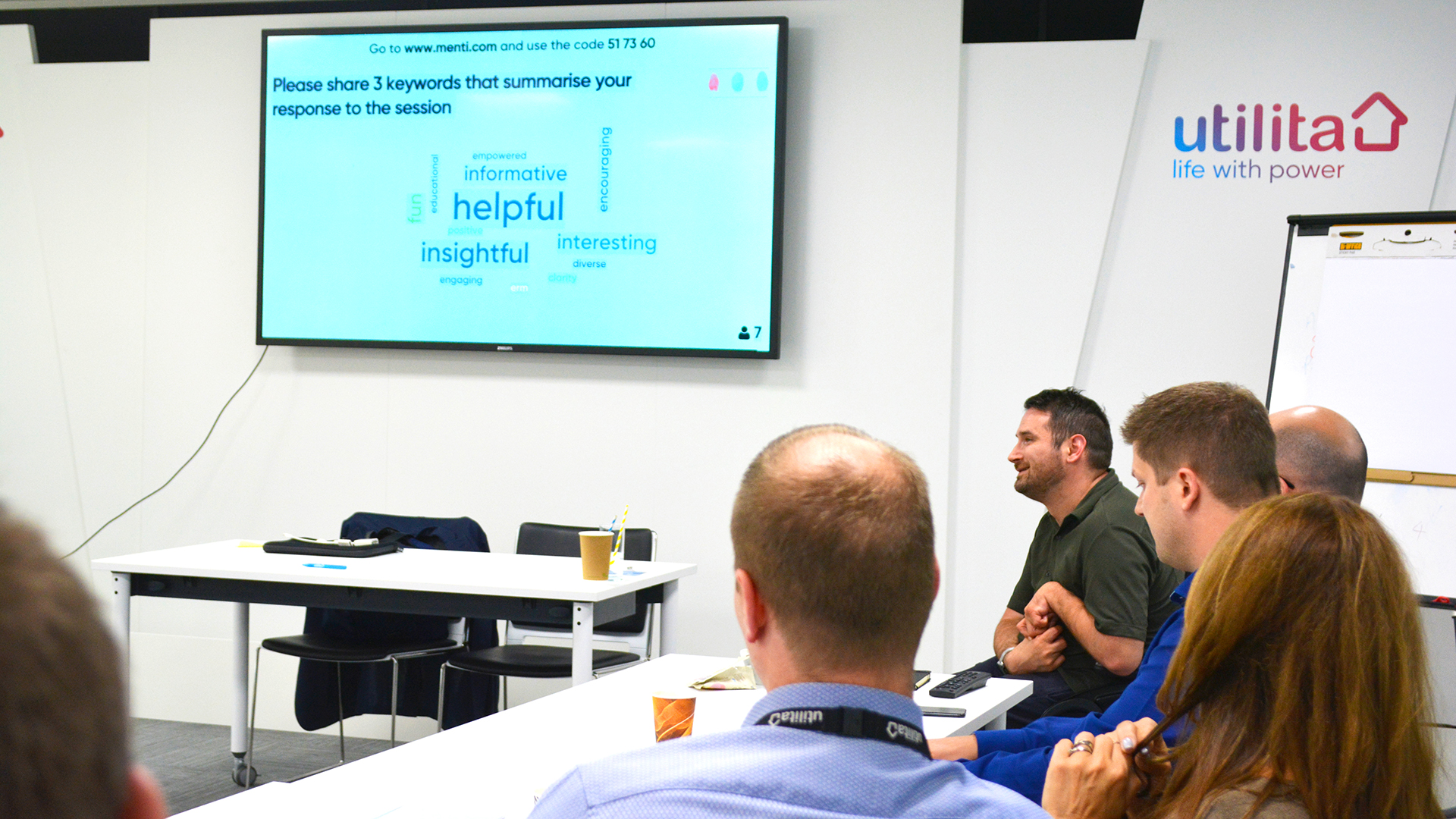 A photo of Adam Hyland (Director of Accessibility and Inclusion) delivering a workshop to the Utilita team. He is talking, sitting in his wheelchair, next to Utilita team members who are sitting at a desk. The powerpoint displays a screen which asks the question 'Please share the 3 keywords that summarise your response to the session.' Below is a wordcloud, where the biggest words are helpful, insightful, informative, interesting and engaging.